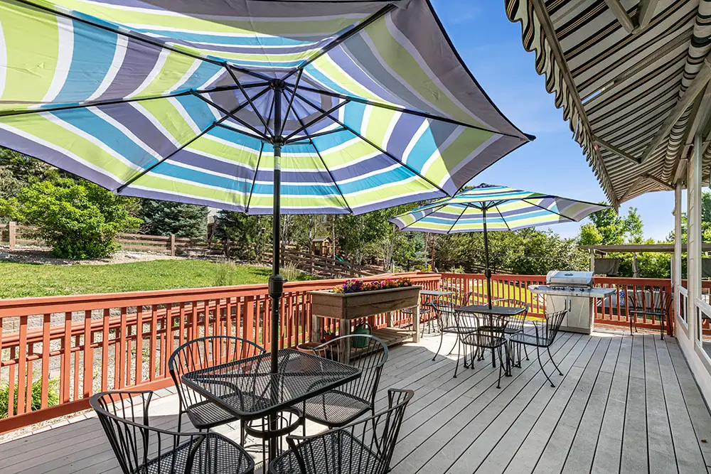 Outdoor wooden deck with black metal tables and chairs, large striped umbrellas, potted flowers, and a barbecue grill, surrounded by greenery under a blue sky.