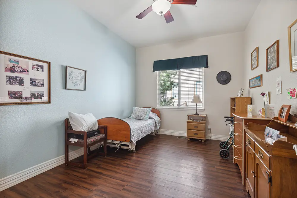 A single bed with wood frame, a chair, a walker, and wooden furniture in a tidy bedroom with photos on the walls, wood floor, and window with blinds.