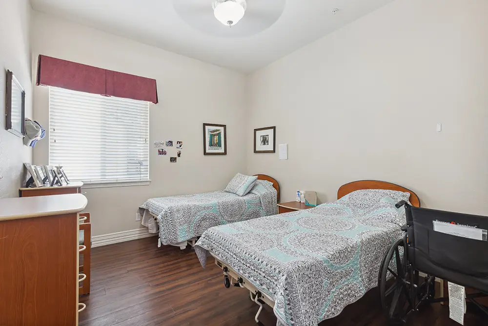 A shared bedroom with two single beds, a wheelchair beside one bed, framed pictures on the walls, and a window with blinds letting in light.
