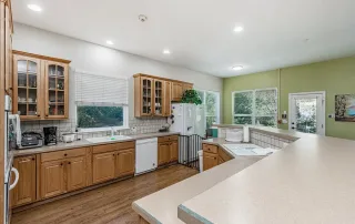 Parker-gallery-10 Spacious kitchen with wooden cabinets, white appliances, large windows with blinds, and green accent wall, featuring ample counter space and natural light.