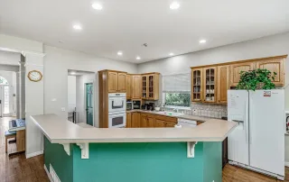 Parker-gallery-11 A kitchen with wood cabinets, white appliances, a green breakfast bar, and recessed ceiling lights. A plant sits on top of the refrigerator near a window with blinds.