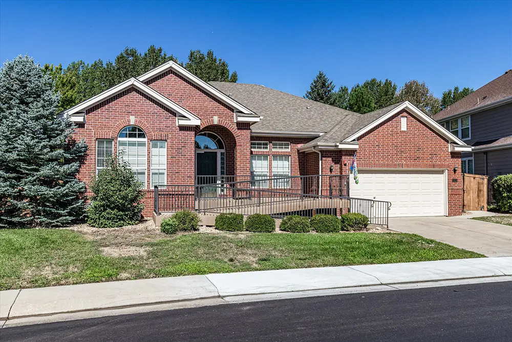 Single-story red brick house with a two-car garage, front ramp for accessibility, manicured shrubs, and a sloped lawn.