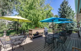 Parker-gallery-4 Outdoor patio with metal tables and chairs, large sun umbrellas in yellow and blue, a raised planter, and a grill, surrounded by trees on a sunny day.