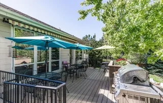 Parker-gallery-5 Outdoor patio with tables, chairs, blue umbrellas, and a barbecue grill, surrounded by greenery and trees under a sunny sky.