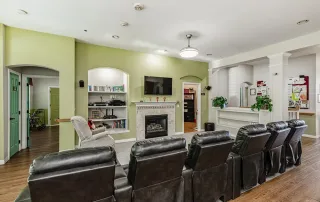 Parker-gallery-7 A row of black recliners faces a wall-mounted TV above a fireplace in a communal living room with green and white walls, bookshelves, and a visible kitchen area.