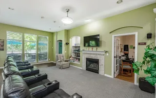 Parker-gallery-8 Room with several recliners facing a TV above a fireplace, large windows, a plant, and an office visible through glass doors on the right.