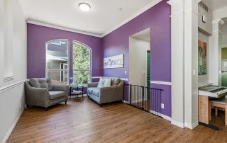 Parker-gallery-9 A small living room with two gray armchairs, a round side table, wood flooring, and purple accent walls. Large window lets in natural light; a safety gate separates the space.