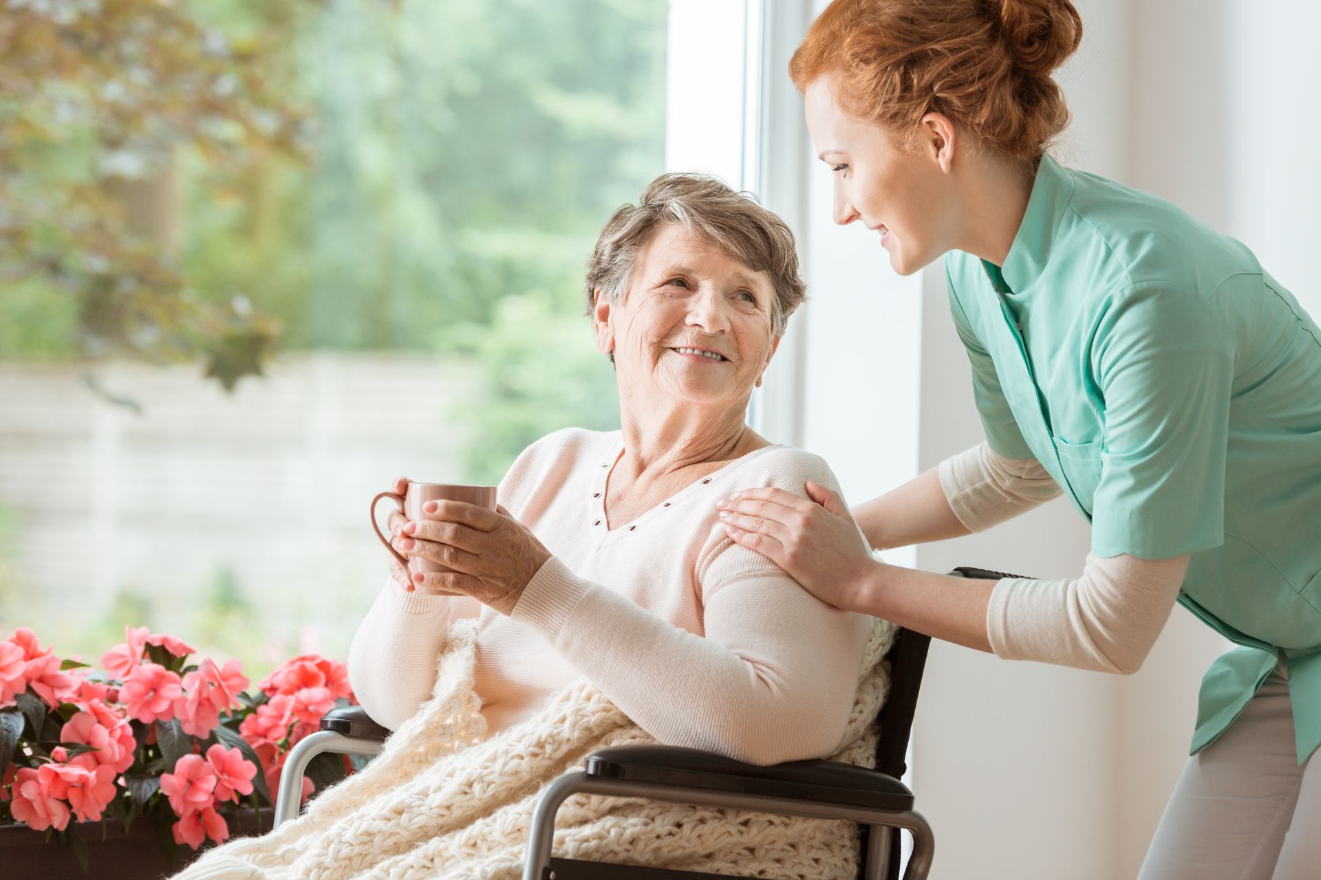 A caregiver in a green uniform smiles and leans toward a senior woman in a wheelchair holding a mug, with pink flowers and a window in the background, creating a safe environment focused on fall prevention.