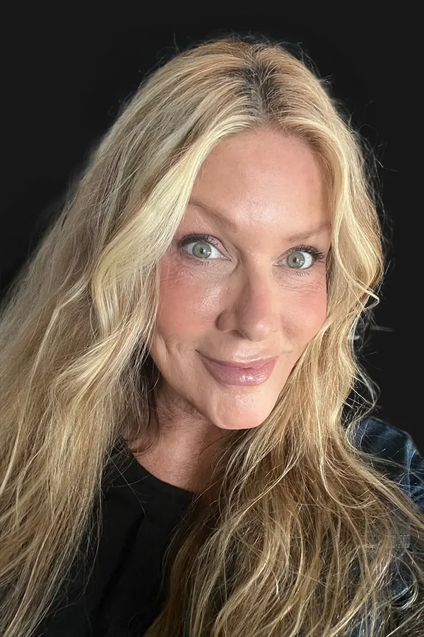 A woman with long, wavy blonde hair and light eyes smiles at the camera against a plain black background.