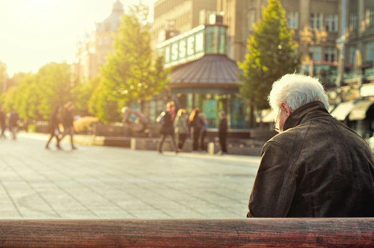 An elderly man with gray hair, possibly experiencing early symptoms of Alzheimer’s, sits alone on a bench in a sunlit urban square as people walk by in the background near a building and trees.