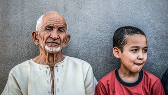 An elderly man with white hair and beard sits next to a young boy in a red shirt against a gray wall, highlighting the bond between generations and the importance of understanding gene variants linked to Alzheimer’s in children and families.