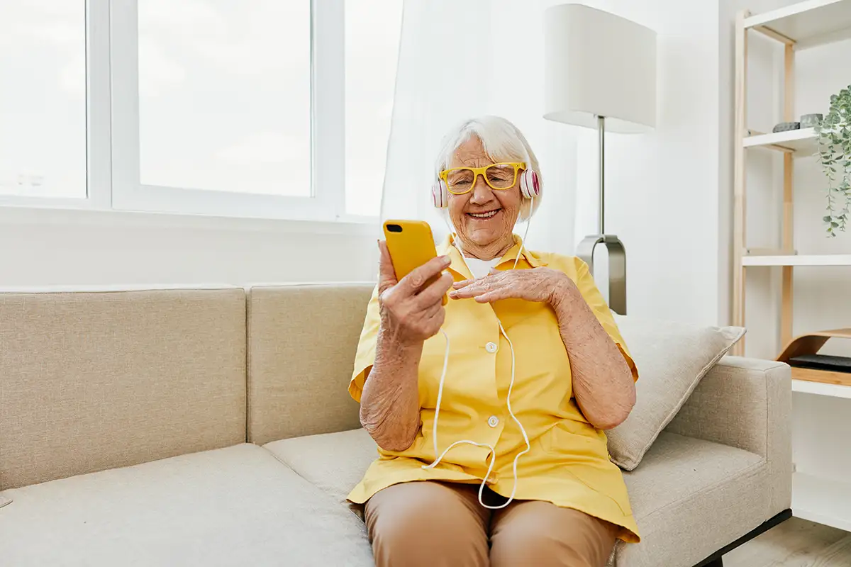 Elderly woman in a yellow shirt and headphones sits on a beige couch, smiling while looking at a yellow smartphone in a bright living room.