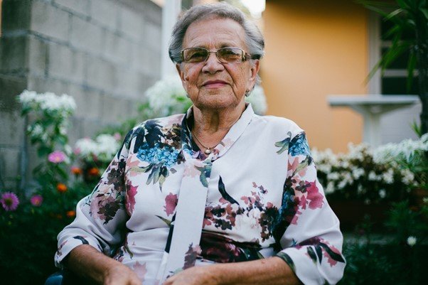 Elderly woman wearing glasses and a floral blouse sits outdoors in a nursing home garden, with flowers and plants visible in the background, highlighting peaceful nursing home aspects.