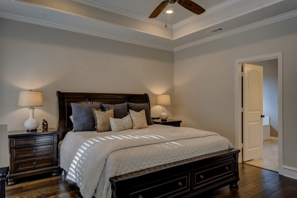 A bedroom with a large dark wood bed, white bedding for a better sleep, two bedside tables with lamps, hardwood floors, and an open door leading to another room.