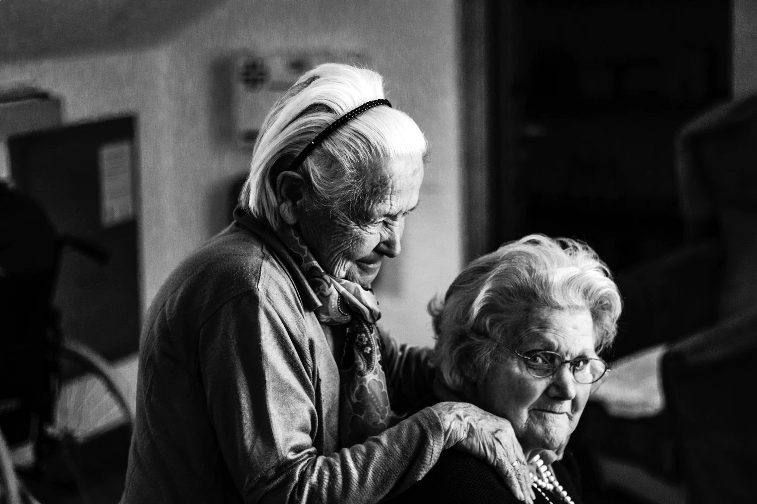Two elderly women in a black-and-white photo; one stands behind the other, gently resting her hands on her shoulders. Both appear calm and thoughtful indoors, reflecting the experiences of many Americans affected by Alzheimer’s.