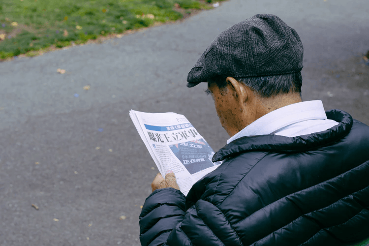 An older person wearing a flat cap and black puffer jacket, possibly affected by low serotonin levels, reads a newspaper while sitting on a park bench near a paved path and grass.