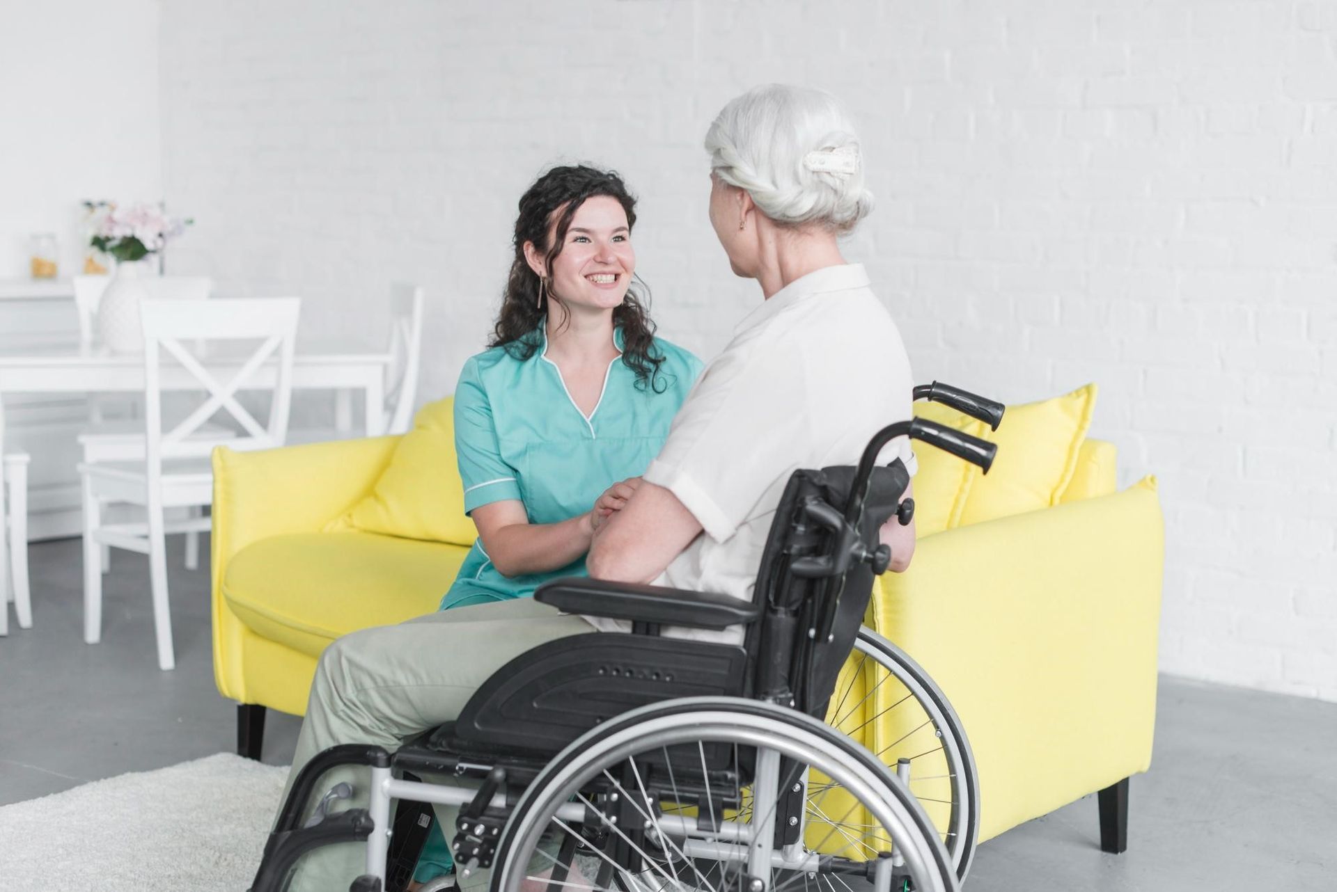 A caregiver in a teal uniform smiles and talks with an older woman in a wheelchair, seated in front of a bright yellow sofa in a light, modern room—highlighting the warmth and dedication of caregivers.