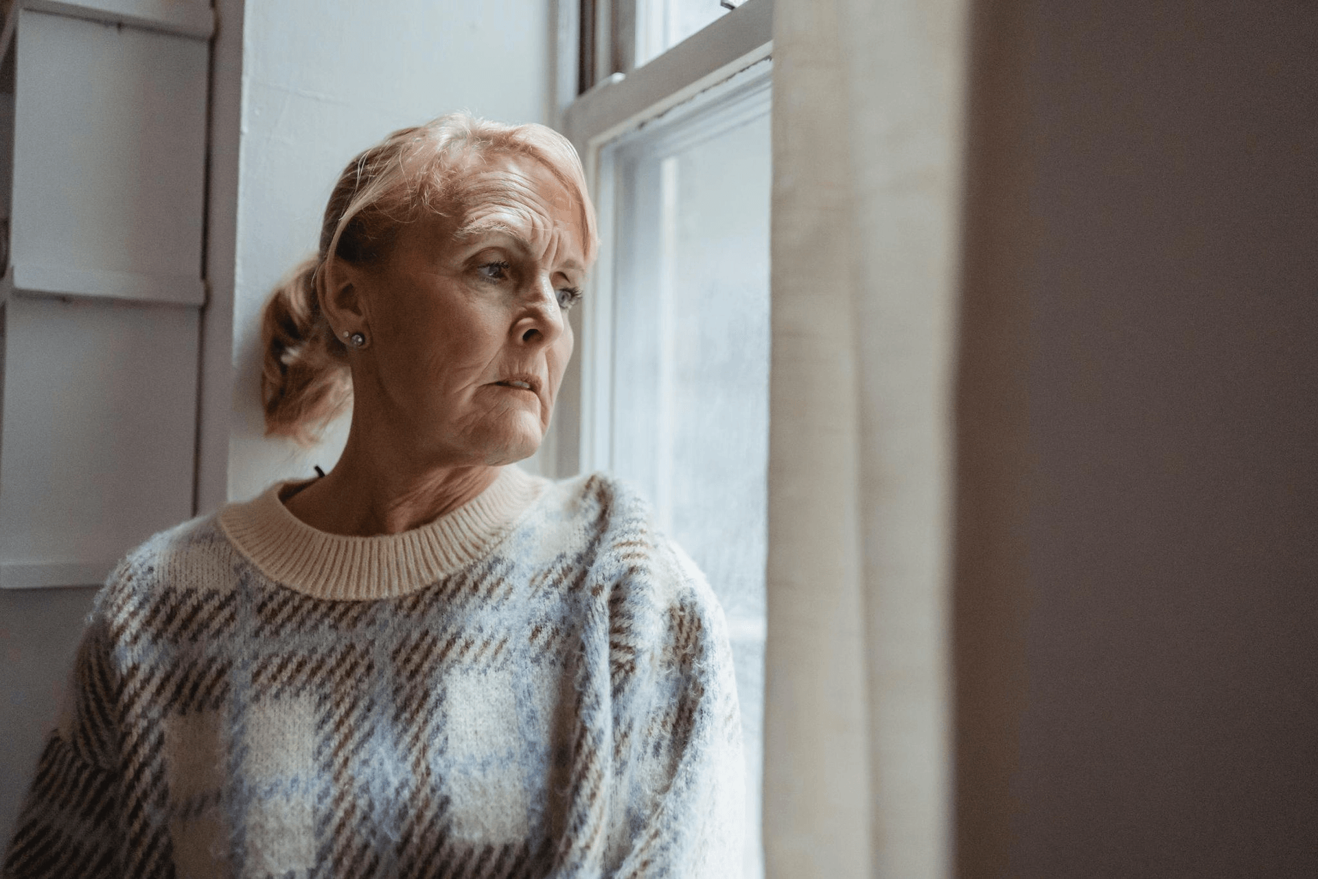 An older woman with light hair in a plaid sweater looks out a window with a neutral expression, sitting indoors by light curtains—a quiet moment sometimes seen in older adults experiencing personality changes associated with Alzheimer’s.