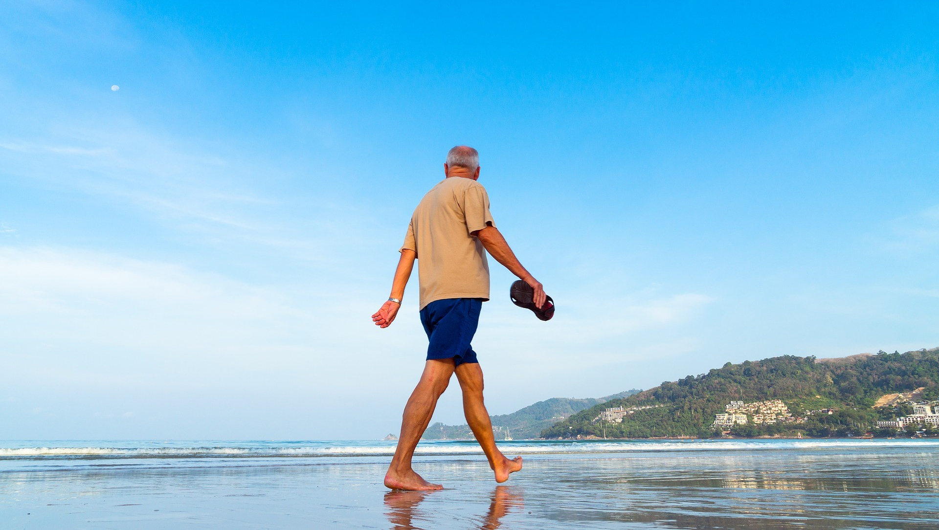 A senior man walks barefoot on a beach holding his sandals, exemplifying wellness habits, with mountains and buildings in the background under a clear blue sky.