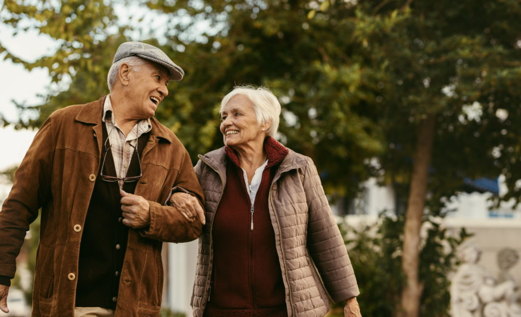 An elderly man and his loved one walk arm in arm outdoors, smiling at each other with trees and greenery in the background.