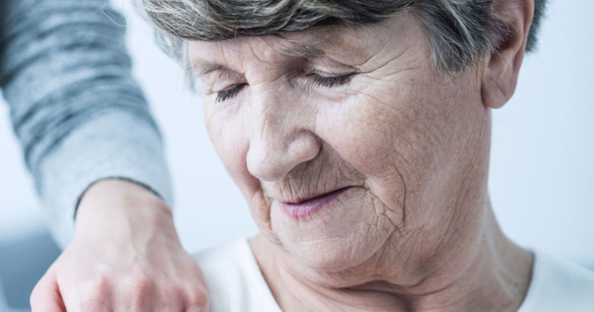 An elderly woman with gray hair, living with Alzheimer’s, smiles gently with her eyes closed while a person’s hand rests reassuringly on her shoulder.