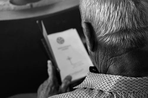 An elderly man with gray hair reads a booklet, seen from behind in black and white. He points to a page on Alzheimer’s risk factors, featuring text and a small emblem at the top.