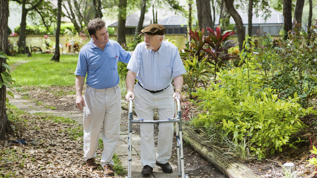 An elderly man using a walker is assisted by a younger man as they walk along a garden path, highlighting the value of budgeting for quality home care cost while enjoying peaceful outdoor moments.