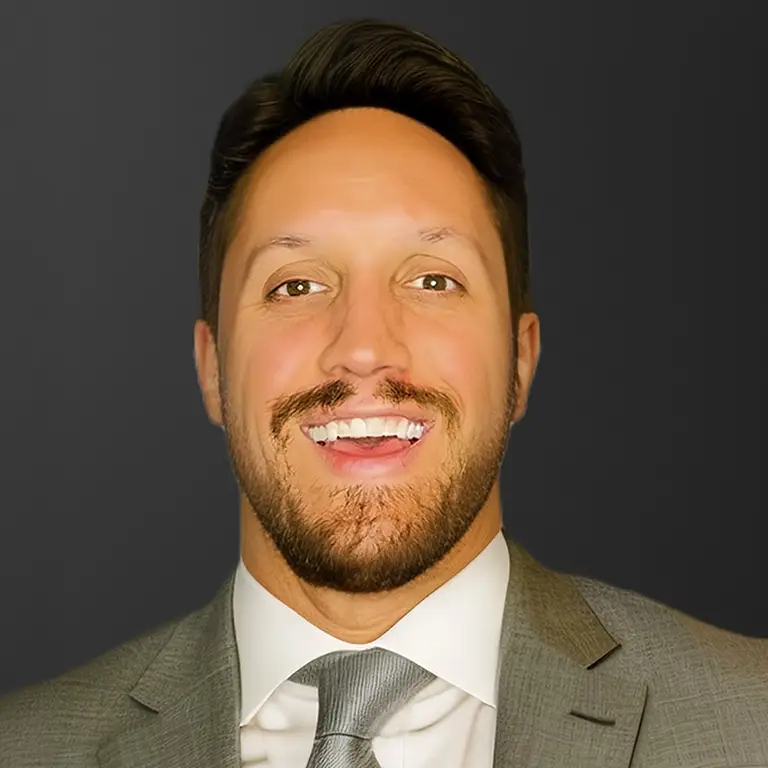 A man in a grey suit and tie smiles at the camera against a plain dark background.