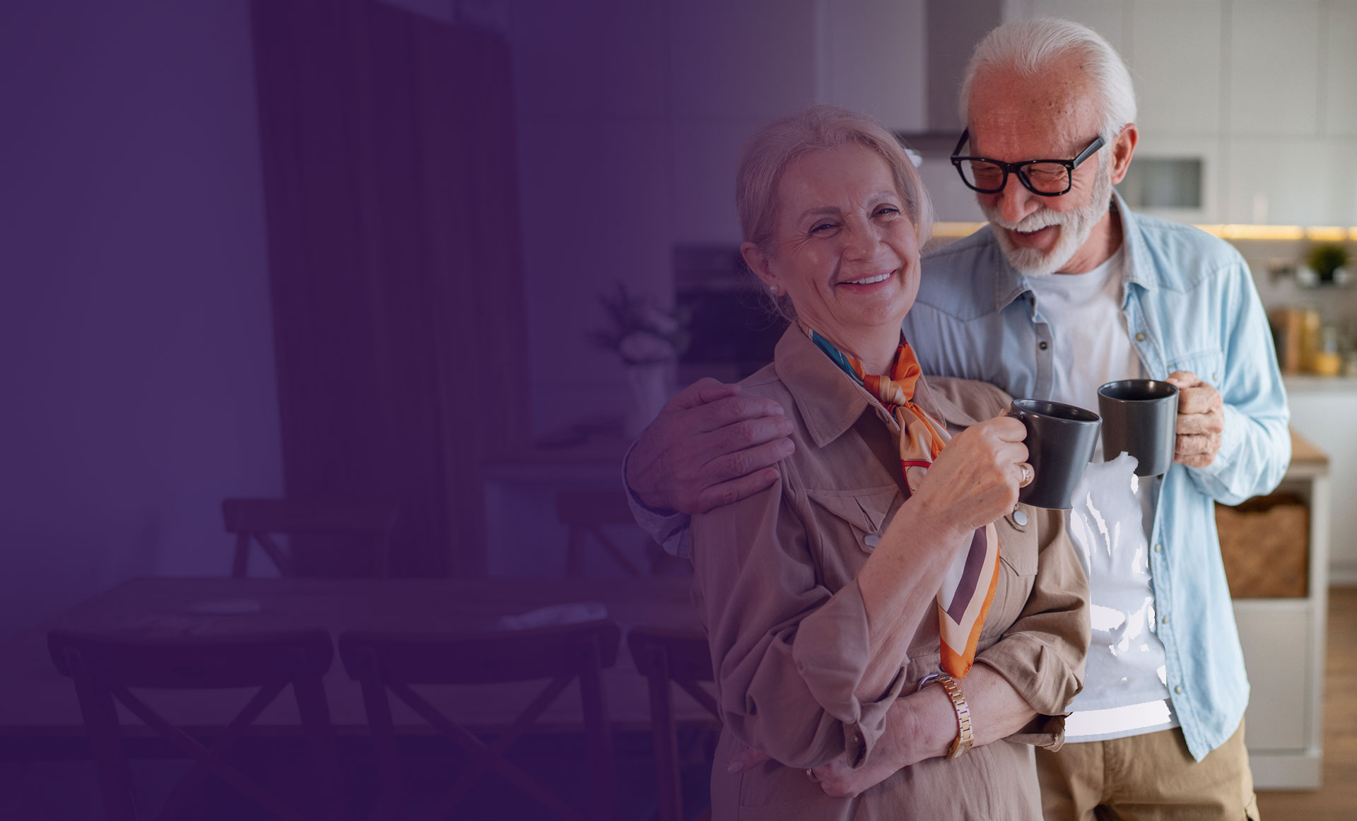 An older couple stands in a kitchen smiling and holding coffee mugs; the man has his arm around the woman.