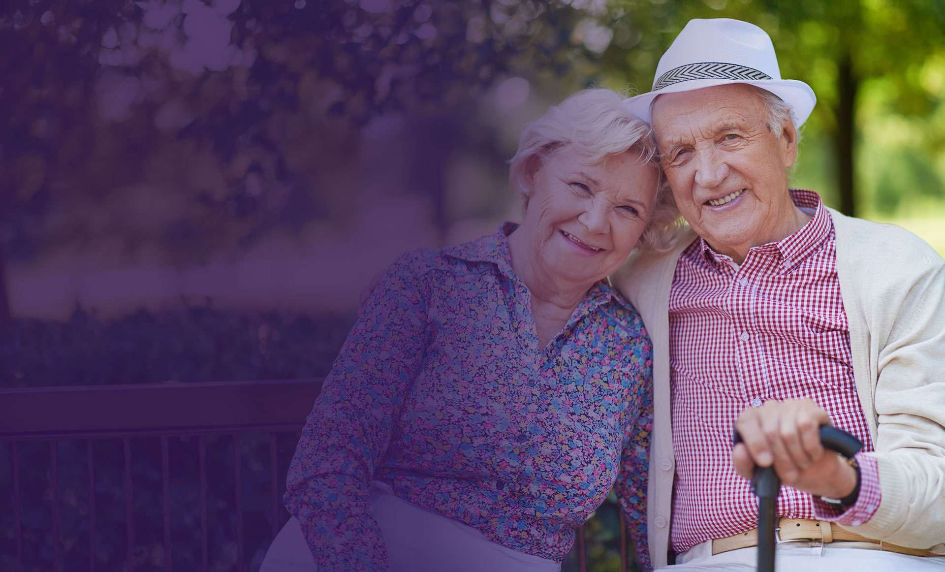An elderly couple sits close together on a bench outdoors, smiling at the camera. The man wears a hat and holds a cane, while the woman rests her head on his shoulder.