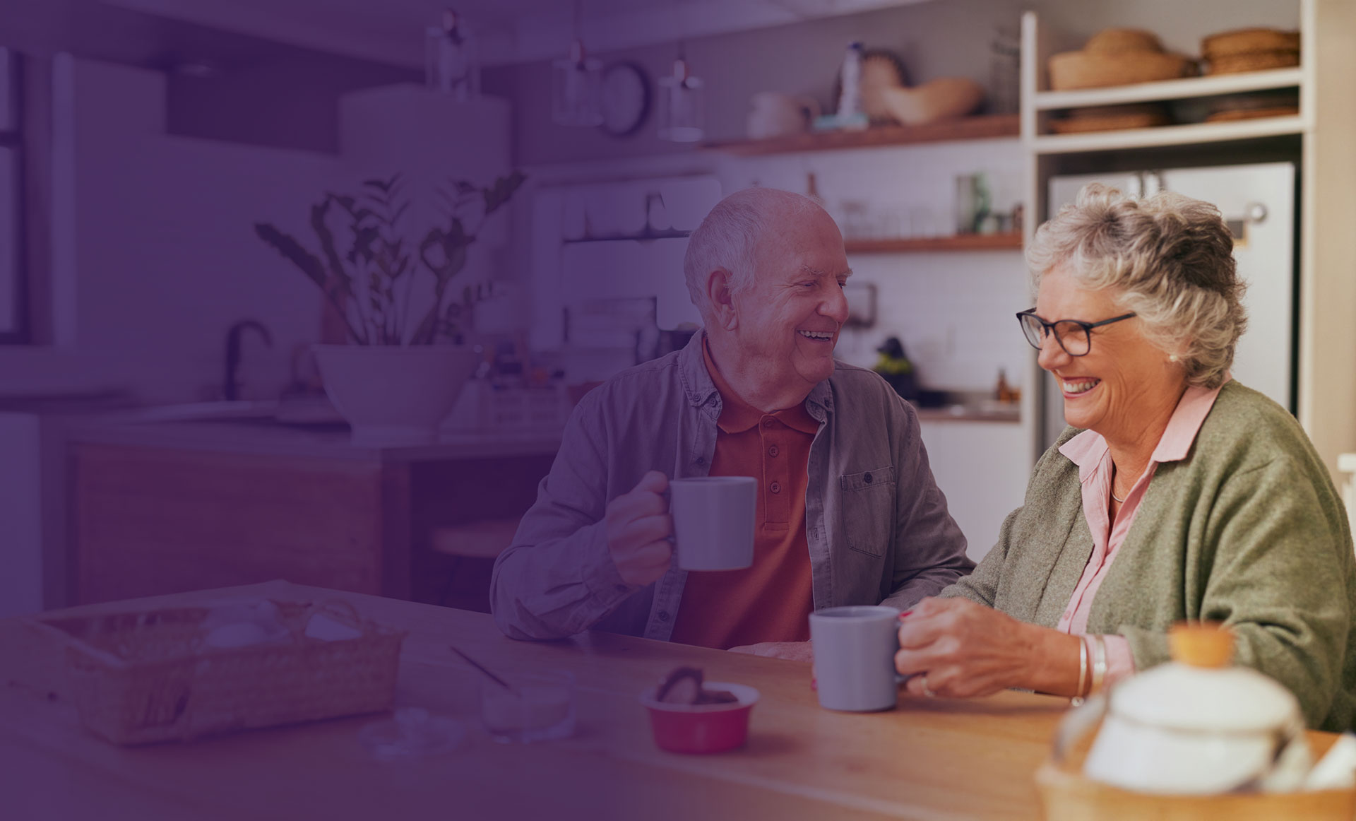 An older man and woman sit at a kitchen table, smiling and holding mugs, with a modern kitchen in the background.