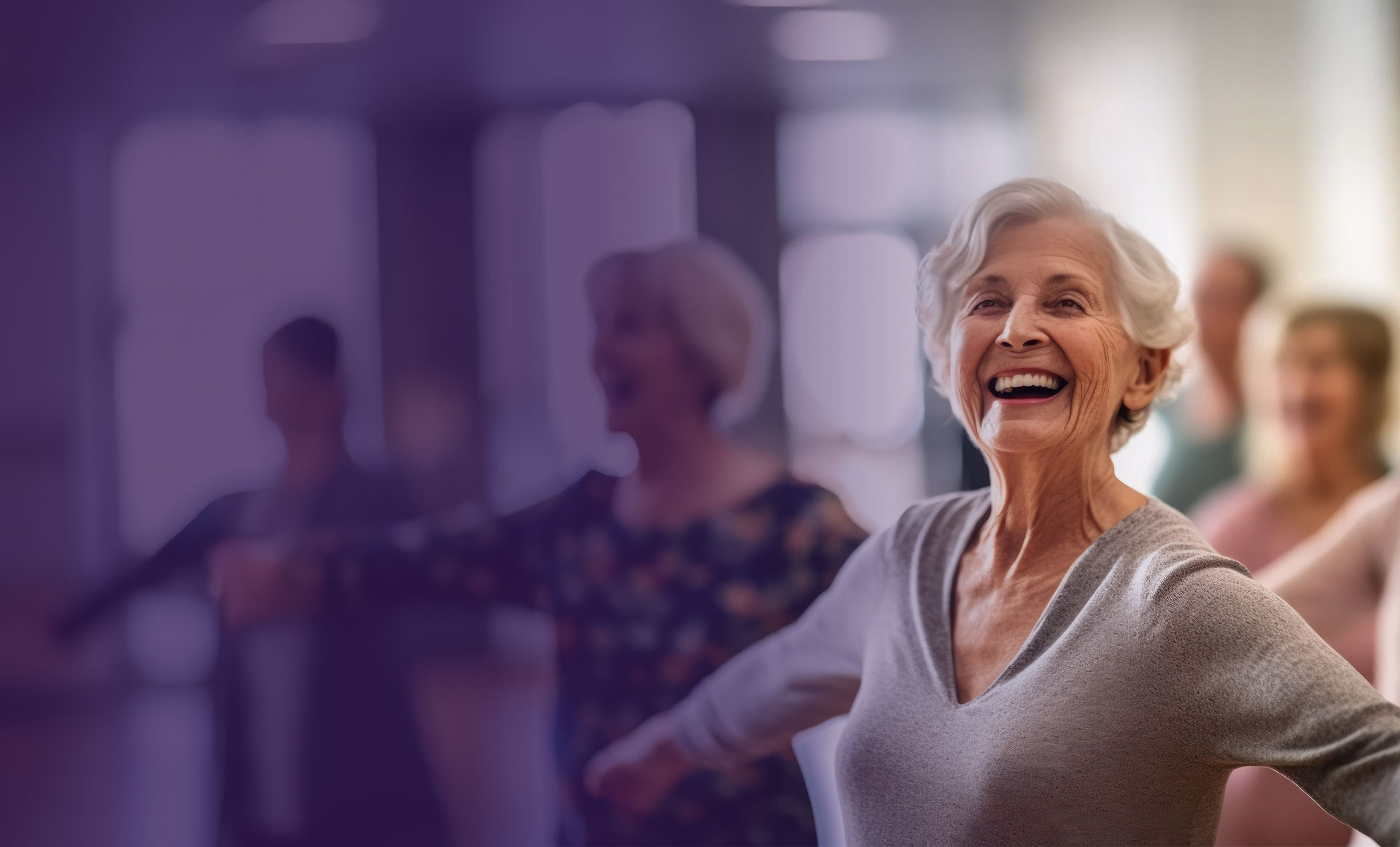 An older woman smiles and stretches her arms in a group exercise class, with other participants blurred in the background.