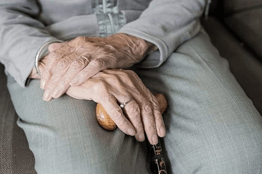 An elderly person’s hands rest on a cane, with visible wrinkles and light-colored clothing, seated on a sofa, perhaps reflecting before having the talk about long-term care.