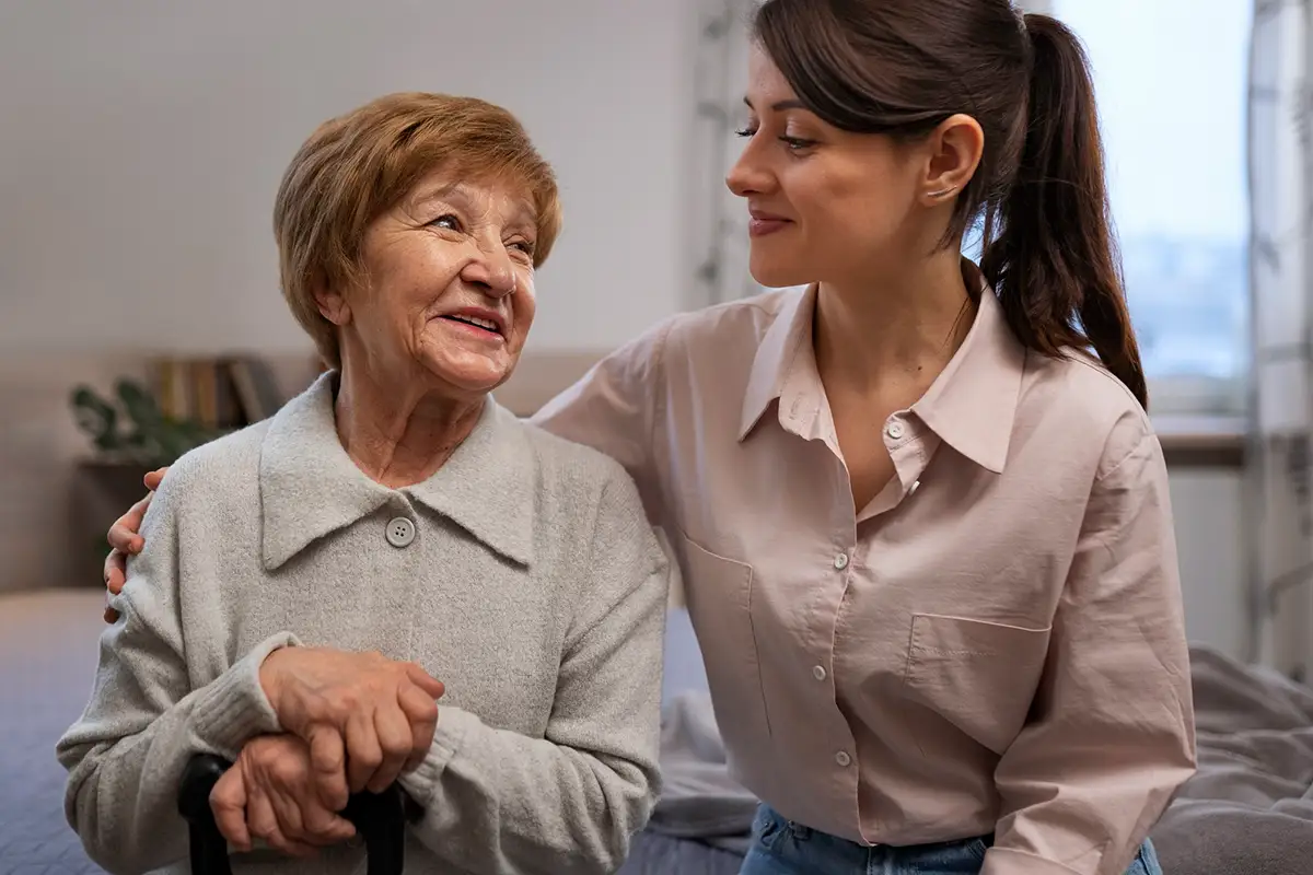 An older woman sits holding a cane while a younger woman, likely her caregiver, places a hand on her shoulder; both are indoors and smiling at each other, reflecting the importance of caregiver mental health in nurturing supportive relationships.