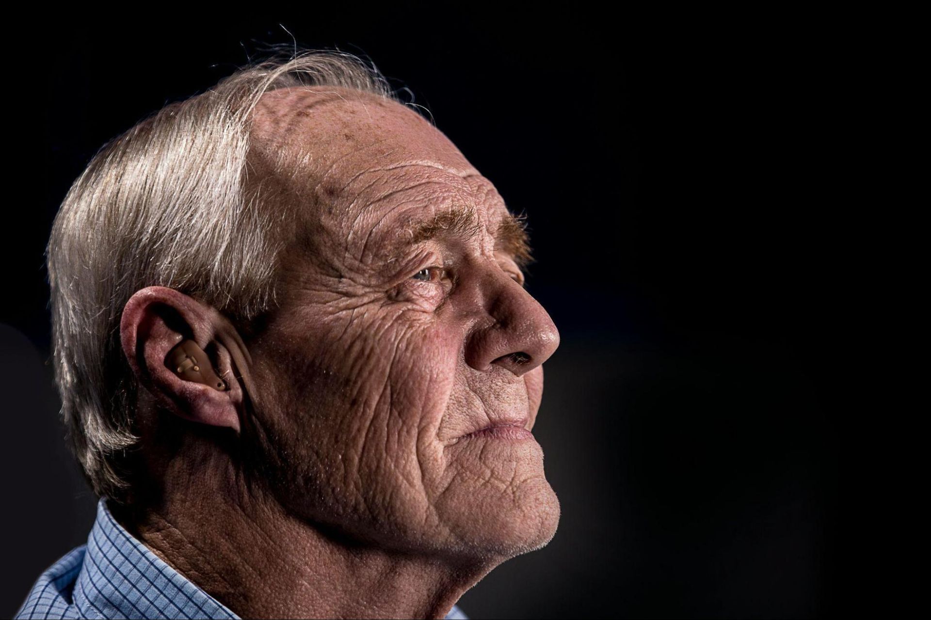 Elderly man with grey hair and a hearing aid gazes thoughtfully to the right, illuminated against a dark background—reflecting on managing legal matters dementia often brings.