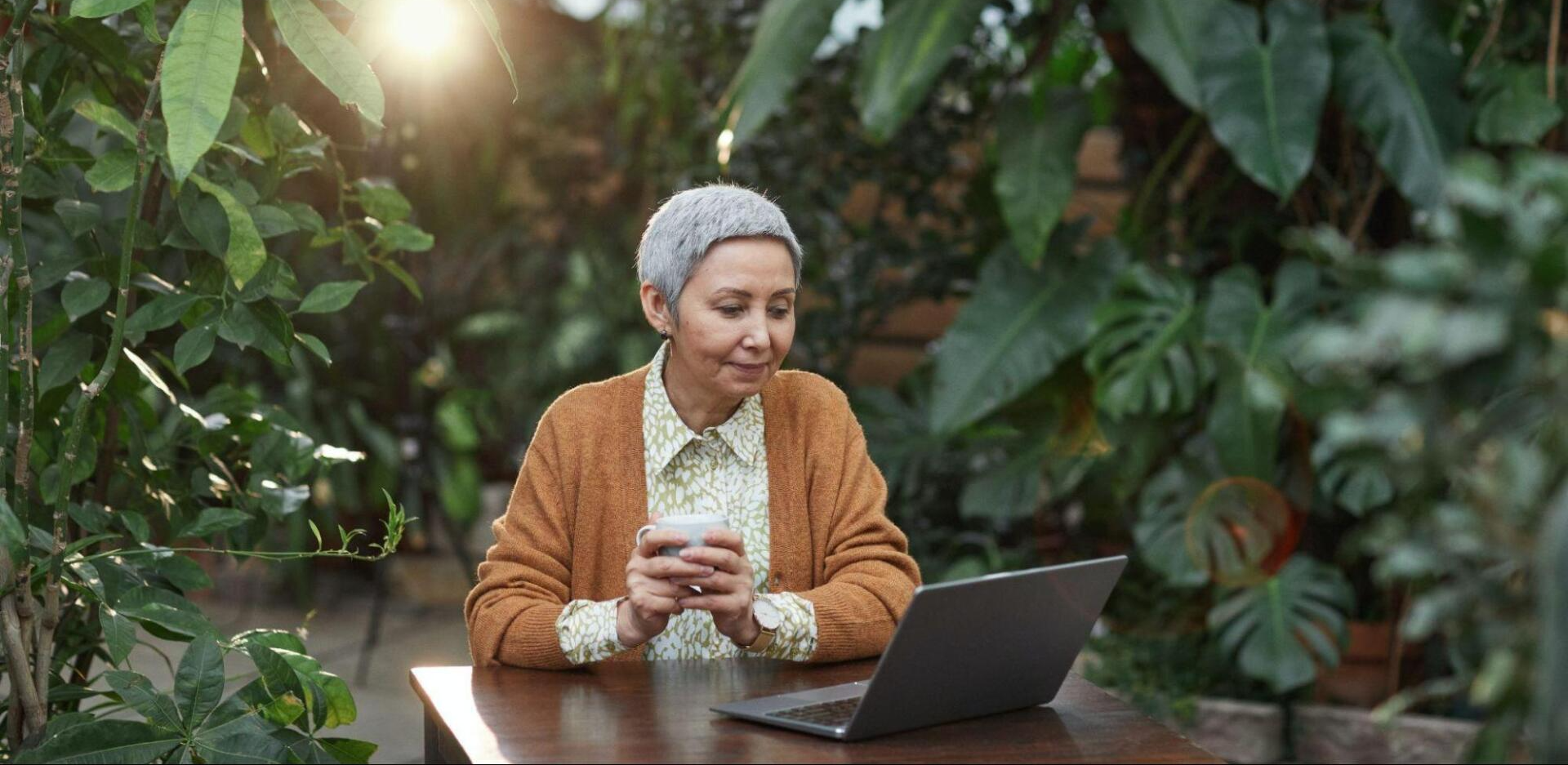 An older woman sits at a wooden table in a garden, holding a cup and looking at a laptop, surrounded by green plants. Sunlight streams in as she explores how seniors can start a business from the comfort of home.