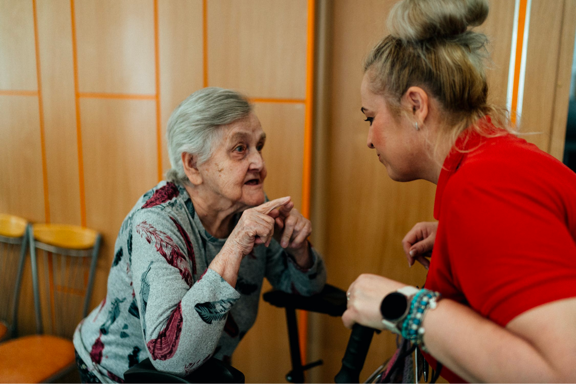 An elderly woman sits and gestures while speaking with a younger woman in a red shirt, who is leaning in to listen—offering guidance for new caregivers on essential self-care.