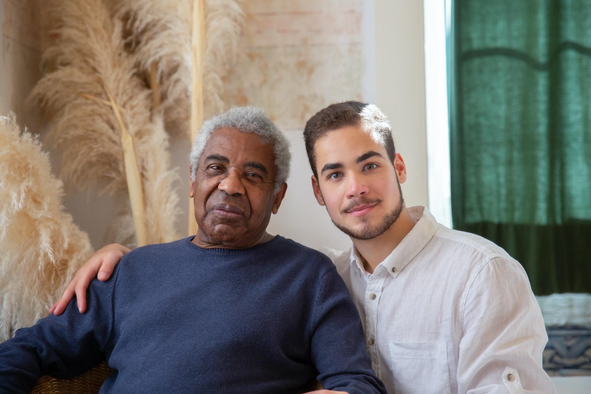 Two men, one older and one younger, sit closely together indoors. The younger man, possibly a senior caregiver, has his arm around the older man. Both are looking at the camera and appear relaxed.