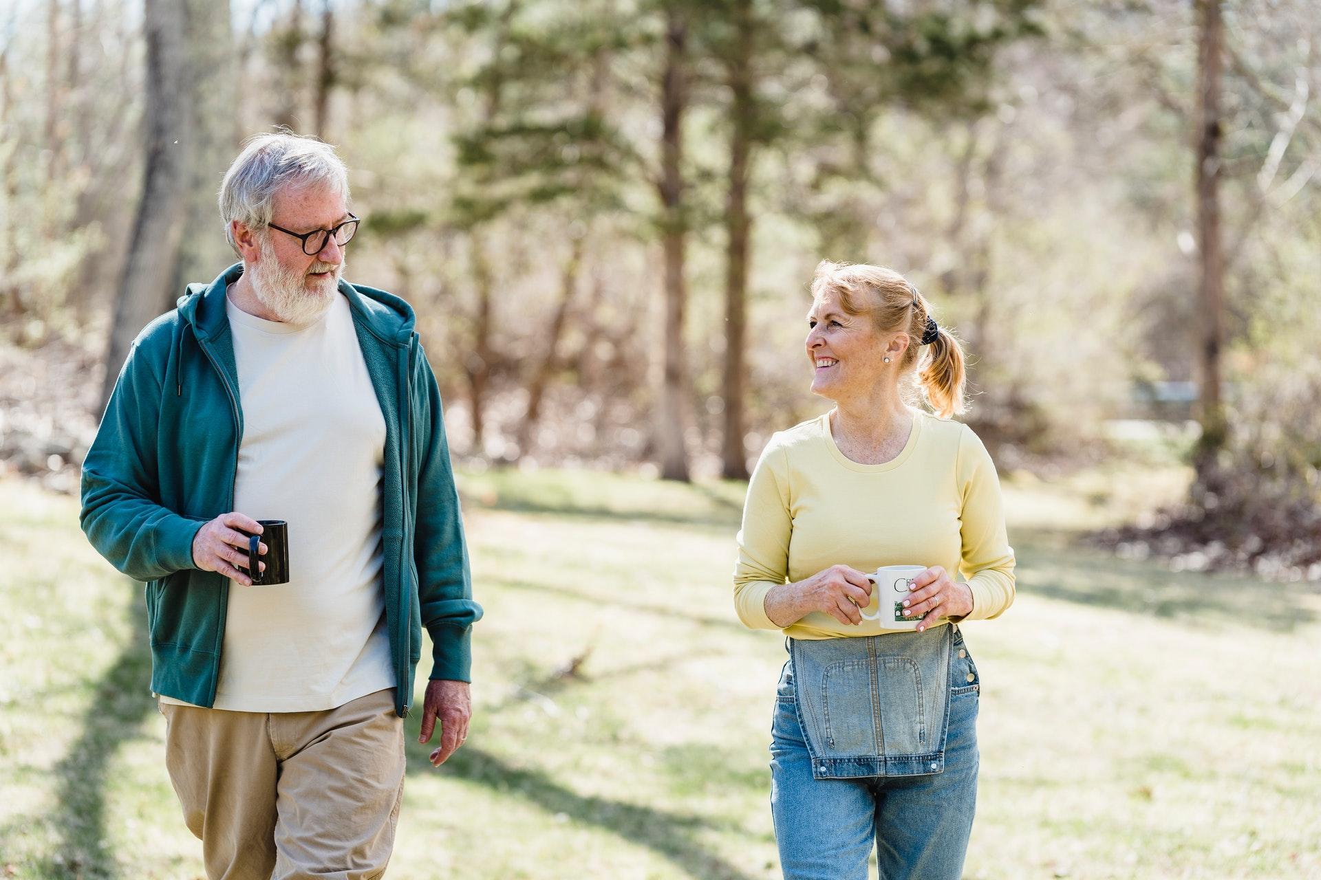 An older man and woman walk outside on grass, holding mugs and smiling at each other, enjoying the sunlight and trees—a heartwarming moment showing how community seniors support each other.