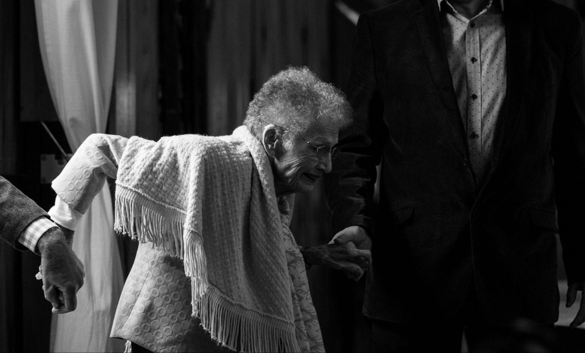 An elderly woman with a shawl is being assisted by two people, one holding her arm and another holding her hand, in a dimly lit setting, highlighting the importance of safety in elderly homes.