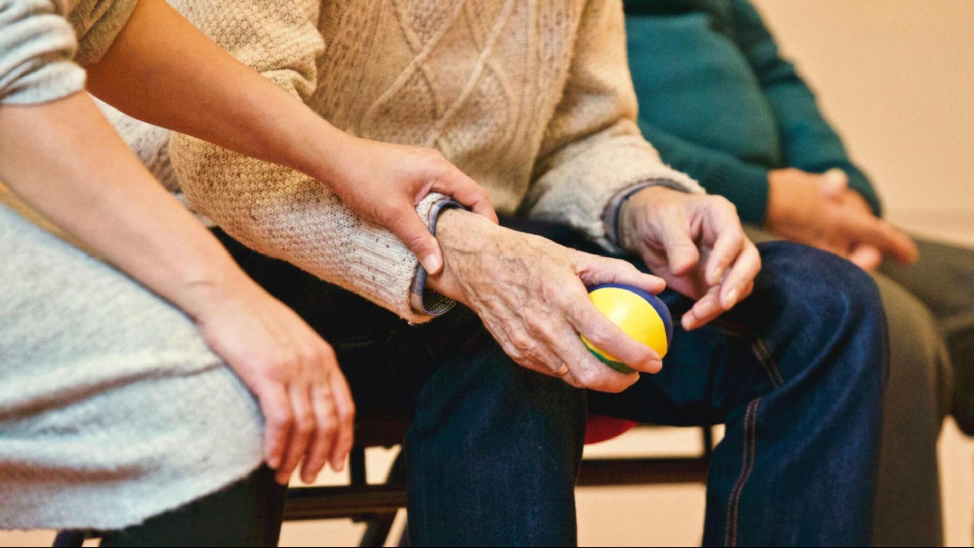A person places a supportive hand on the arm of an older adult holding a yellow and blue ball, with another person seated nearby.