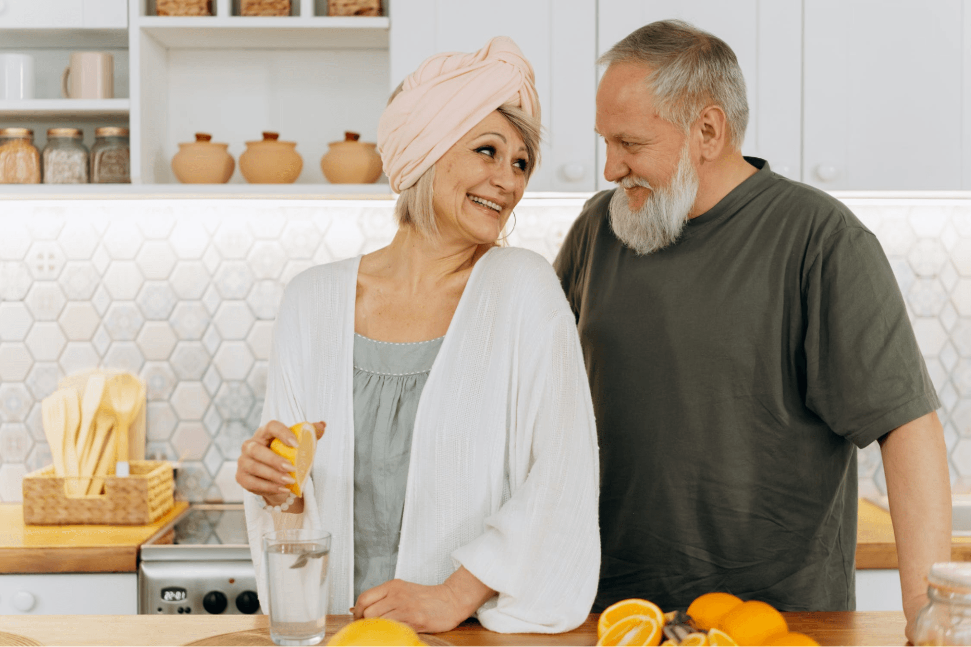 An older woman squeezes a lemon into a glass of water while smiling at an older man in a kitchen with oranges on the counter, enjoying the simplicity that comes with downsizing for seniors.