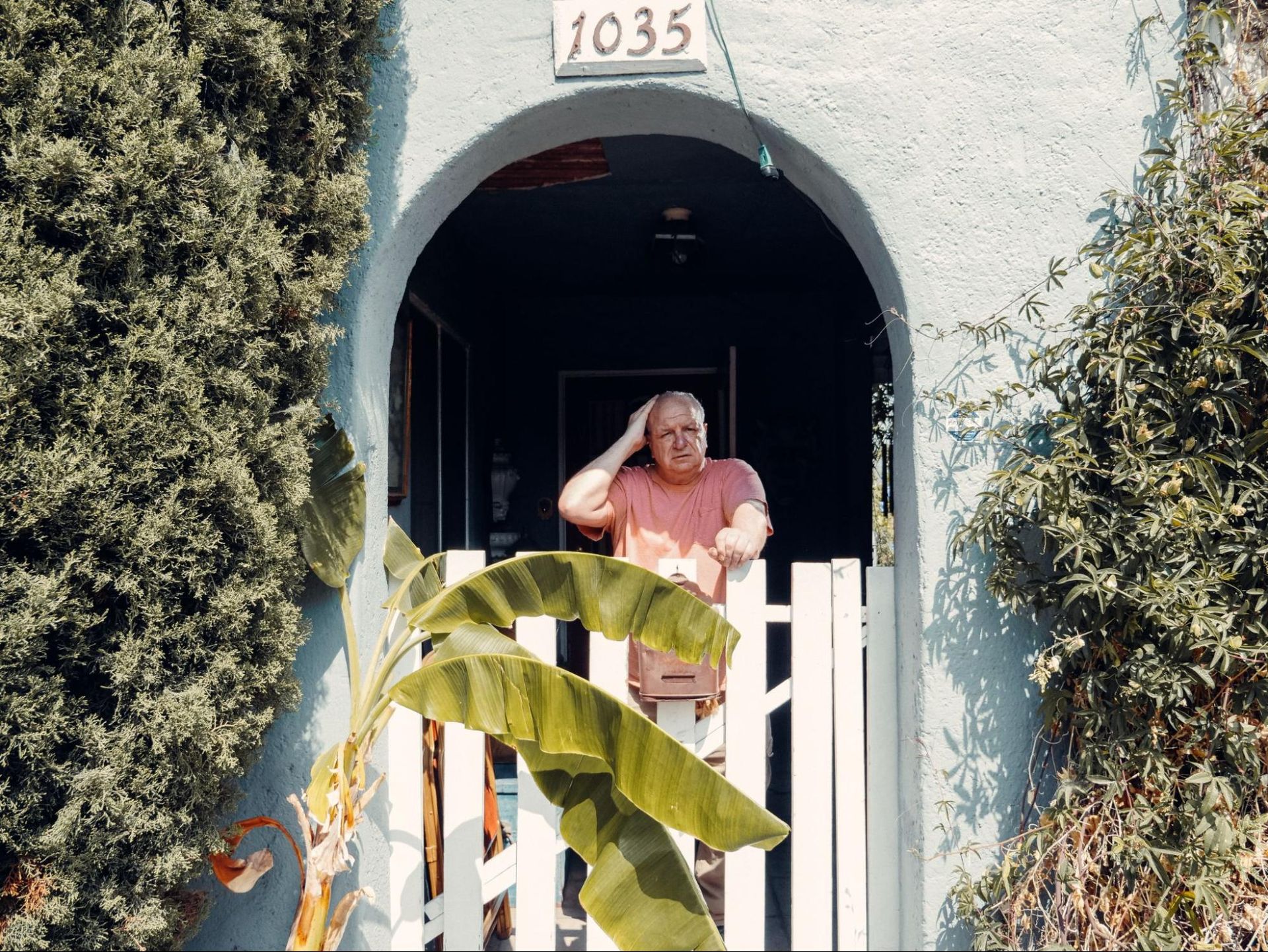 An older man in a pink shirt, living with dementia, stands behind a white gate at the entrance of house 1035, surrounded by greenery and plants.