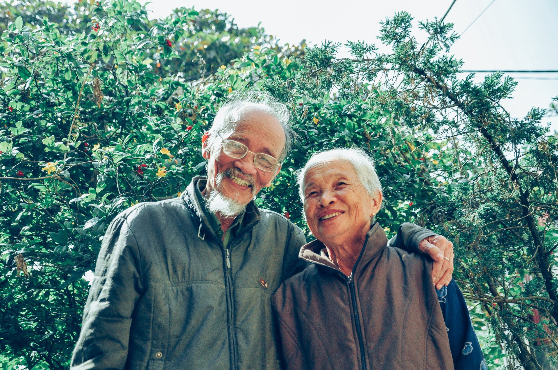 Two elderly people stand close together outside an assisted living community, smiling at the camera, with greenery and sunlight in the background.