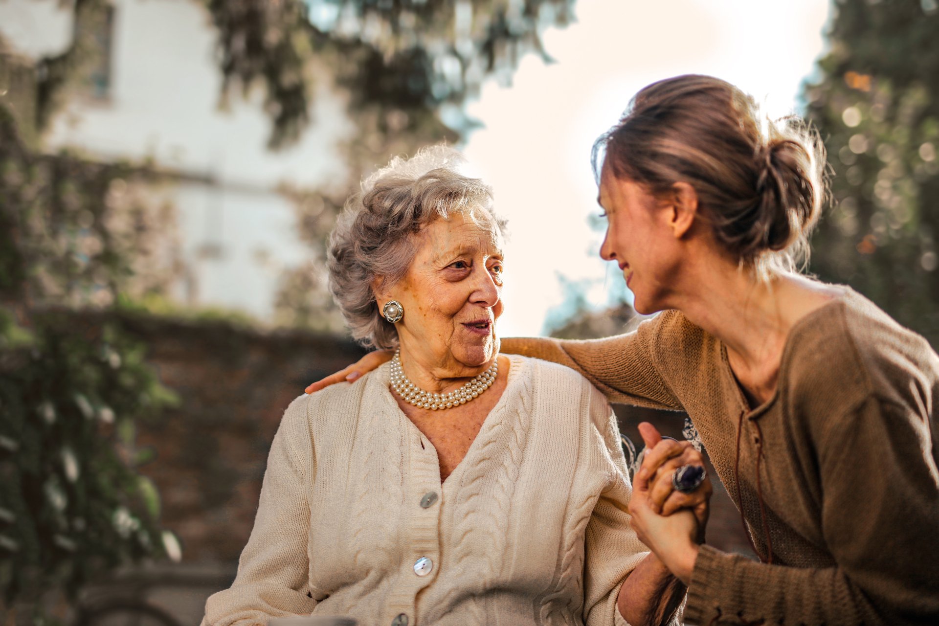 An elderly woman and a younger woman sit together outdoors, smiling and holding hands, sharing a warm moment. This touching scene highlights the importance of connection for seniors, especially those facing challenges like Alzheimer's.