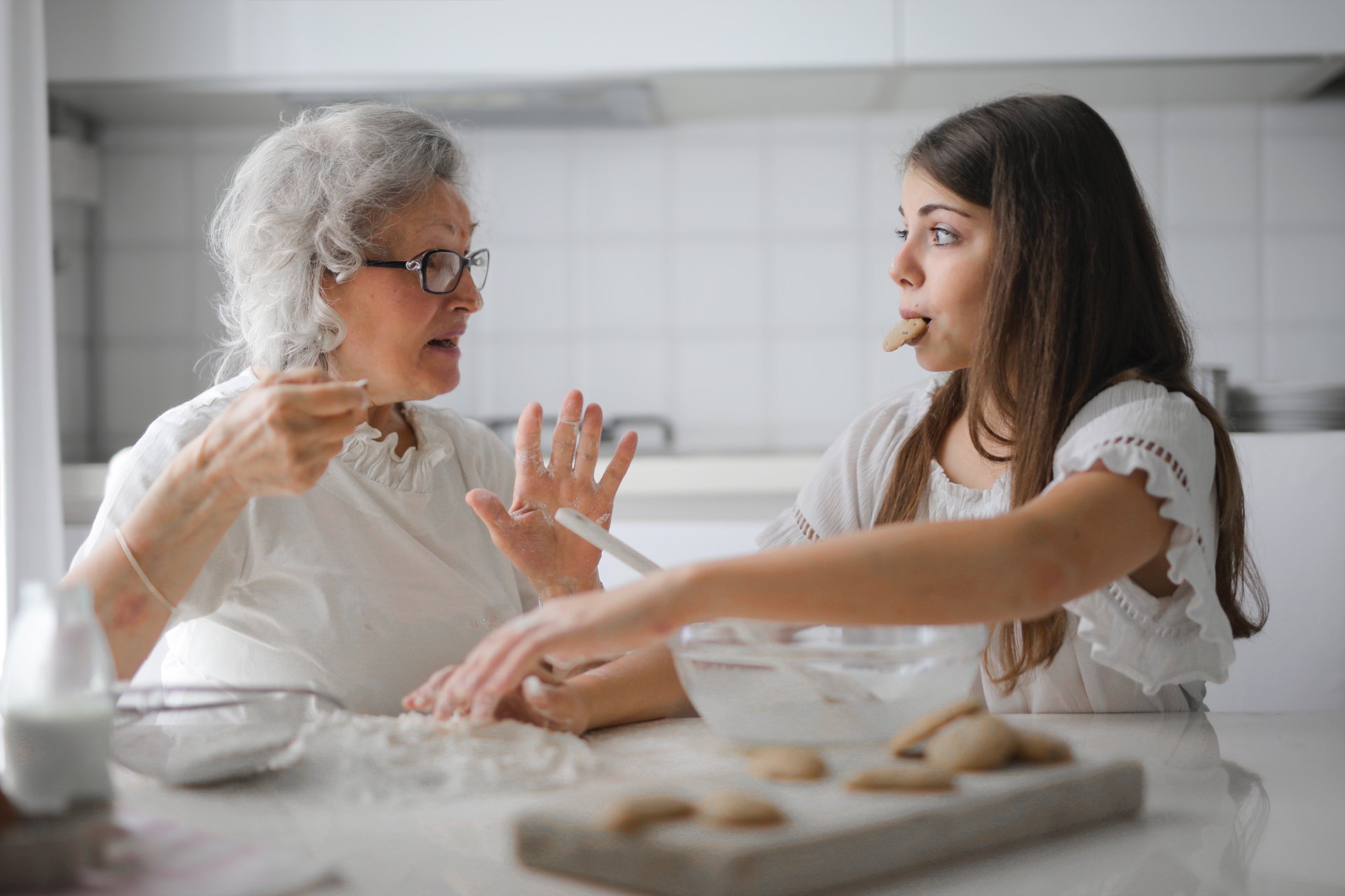 An older woman gestures while talking to a younger woman, who is eating a cookie in a kitchen with baking ingredients—perhaps discussing moving closer to aging parents or caring for elderly parents together.