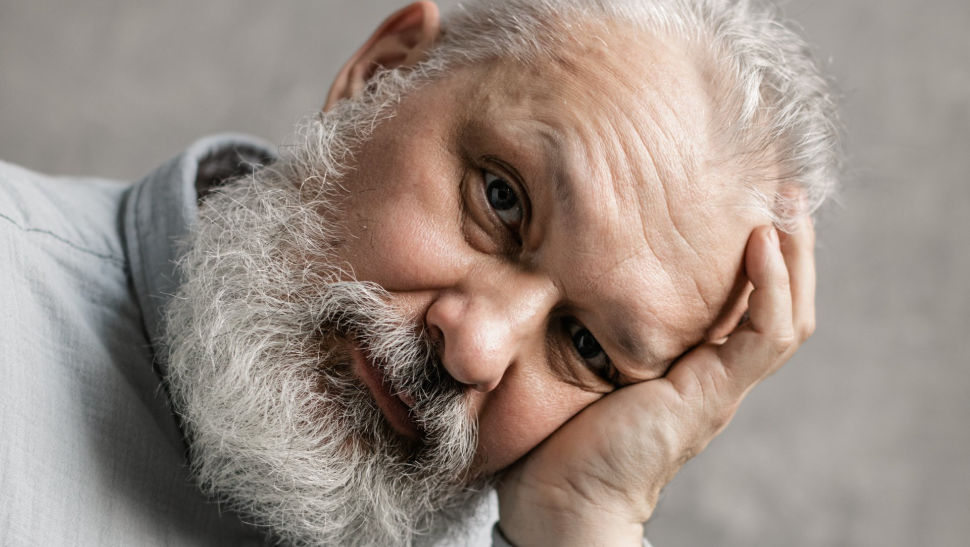 Elderly man with a gray beard rests his head on his hand and looks directly at the camera, wearing a light gray shirt, reflecting the thoughtful moments often seen in Alzheimer Care.