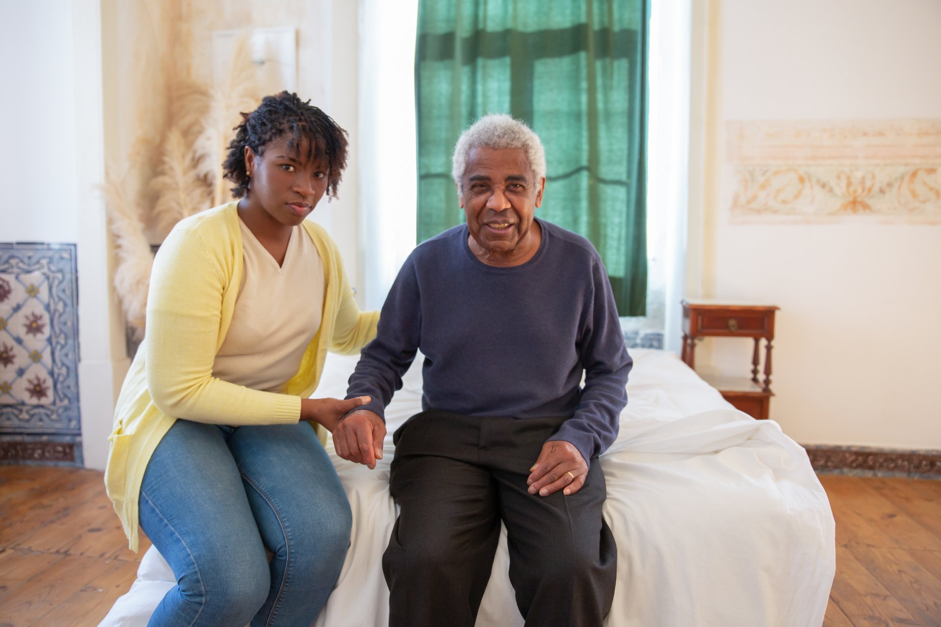 A young woman provides personal care as she sits next to an older man on a bed, holding his hand and looking at the camera in a well-lit room.