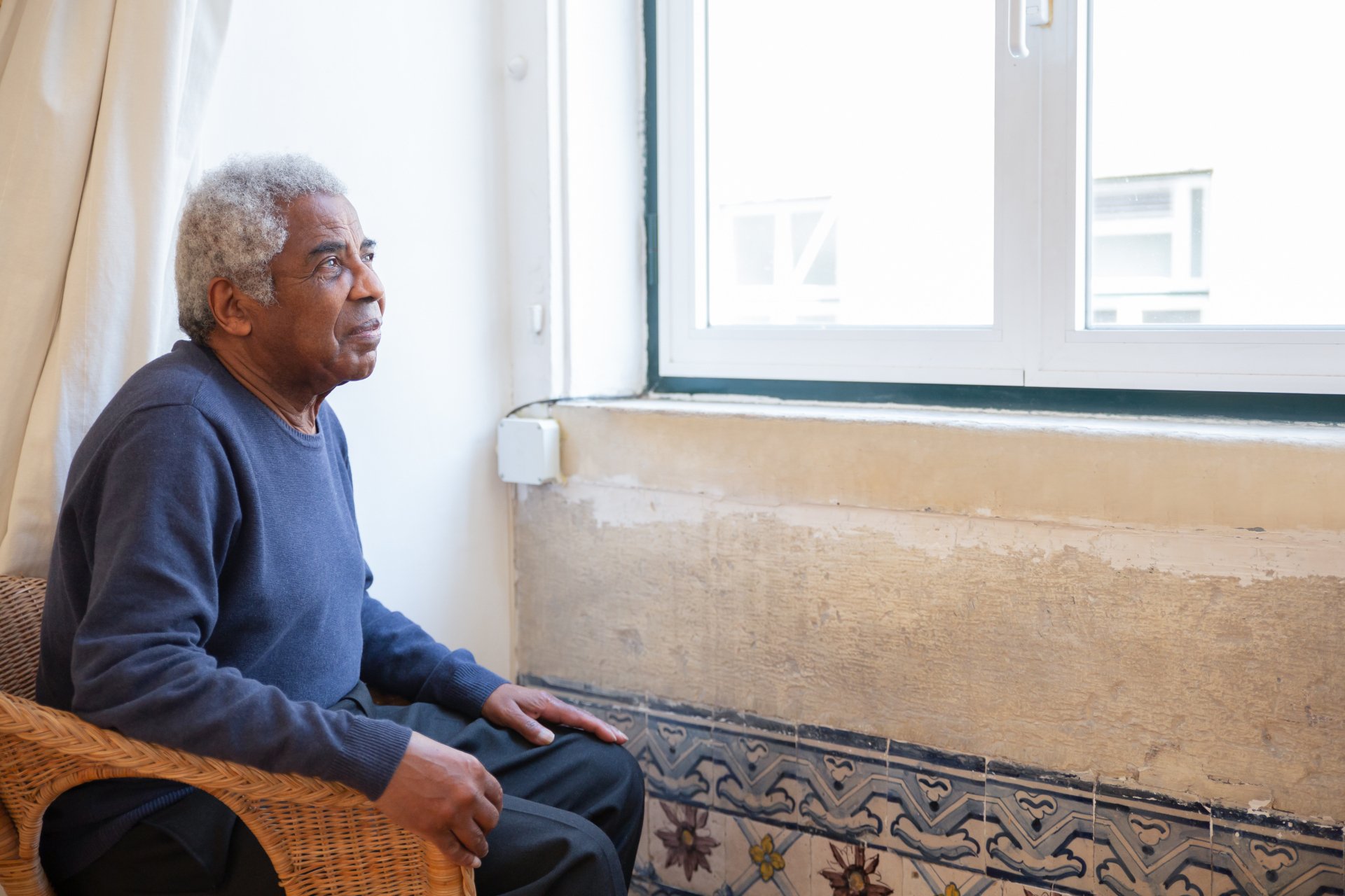 An older man with gray hair sits in a wicker chair indoors, looking out of a window with a neutral expression, considering better options and nursing home alternatives for his loved one.