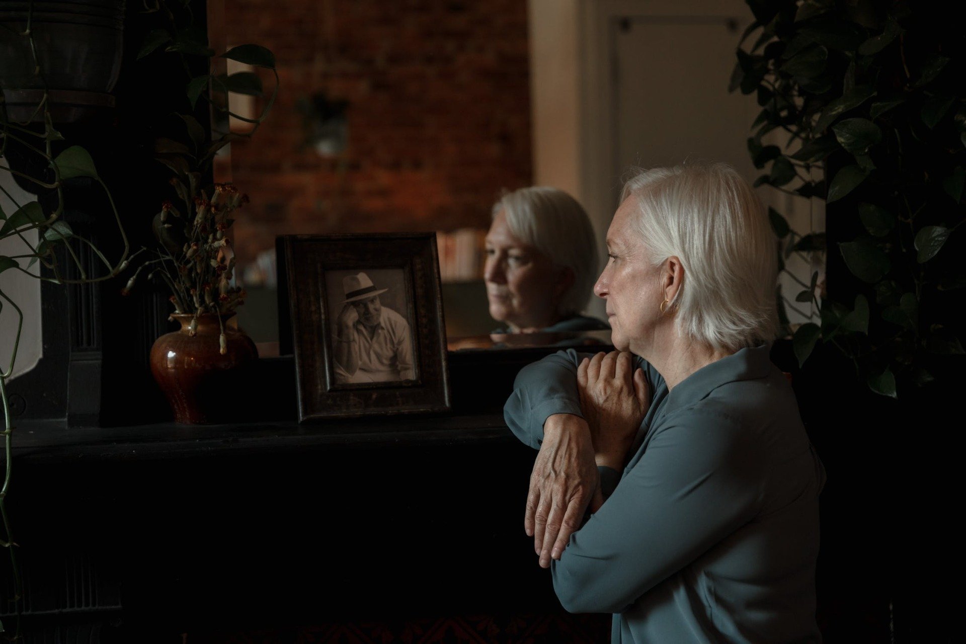 An older woman with white hair sits by a dark mantelpiece, reflecting in a mirror beside a framed photograph of a man and a small vase with dried branches, her calm agitation hinting at the silent struggles of Alzheimer’s disease.